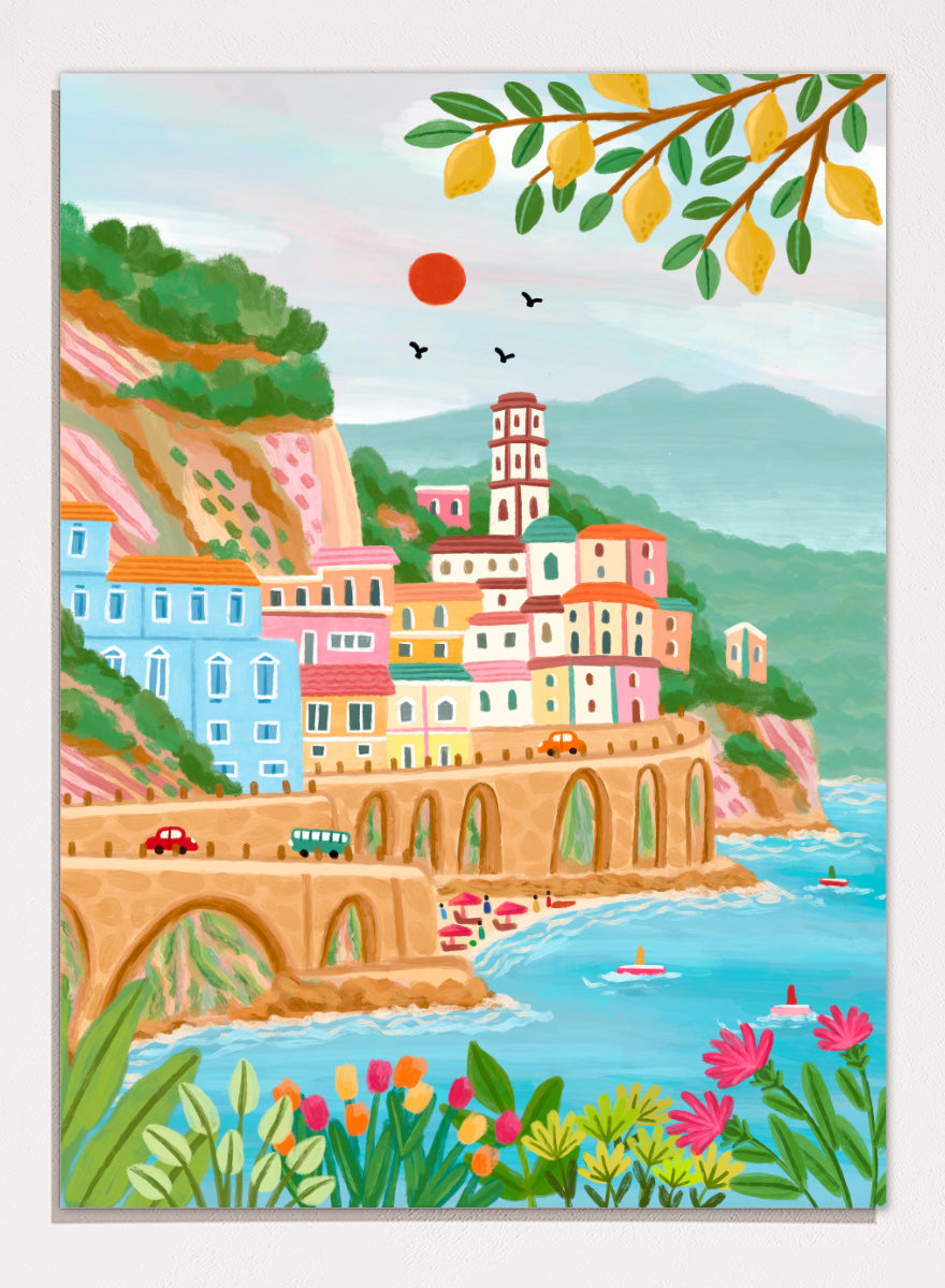 A colorful illustrated coastal town scene with pastel buildings, a stone bridge, blue water, mountains, flowers, and hanging yellow leaves. Shop Colorful Amalfi Coast by Nidhi Kachhadiya, a vibrant Italian coastal wall art print inspired by the Amalfi Coast, pastel villages, blue sea, and lemon trees.