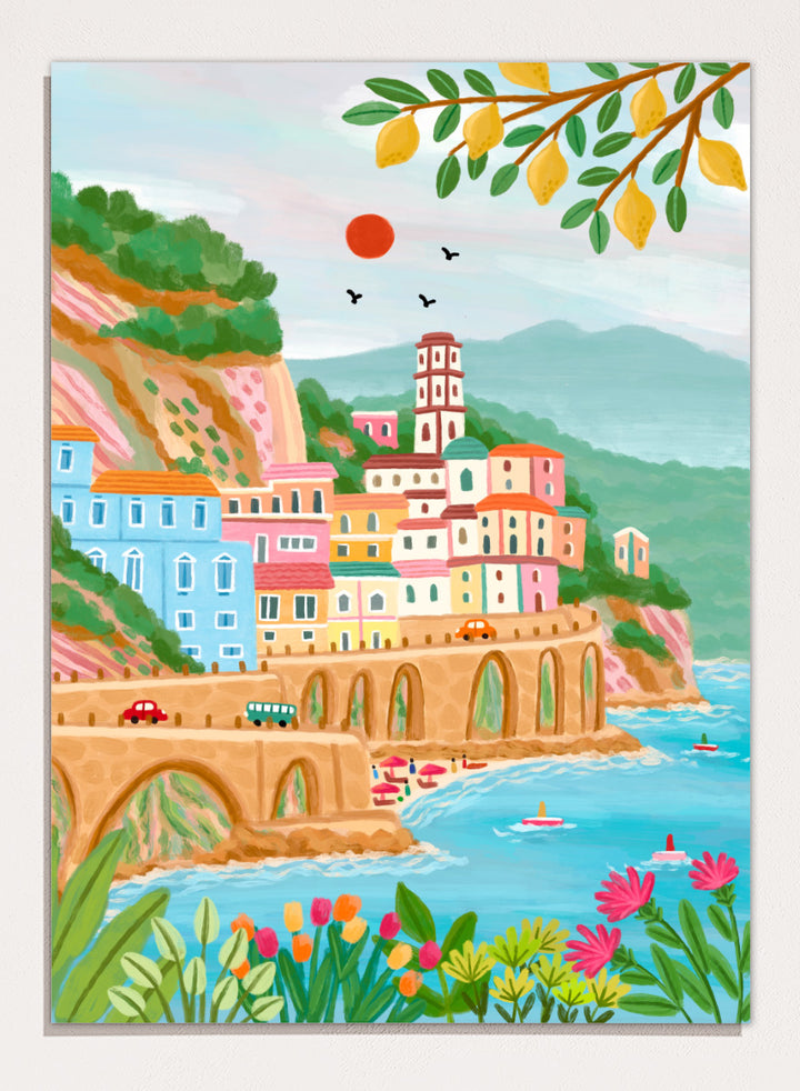 A colorful illustrated coastal town scene with pastel buildings, a stone bridge, blue water, mountains, flowers, and hanging yellow leaves. Shop Colorful Amalfi Coast by Nidhi Kachhadiya, a vibrant Italian coastal wall art print inspired by the Amalfi Coast, pastel villages, blue sea, and lemon trees.