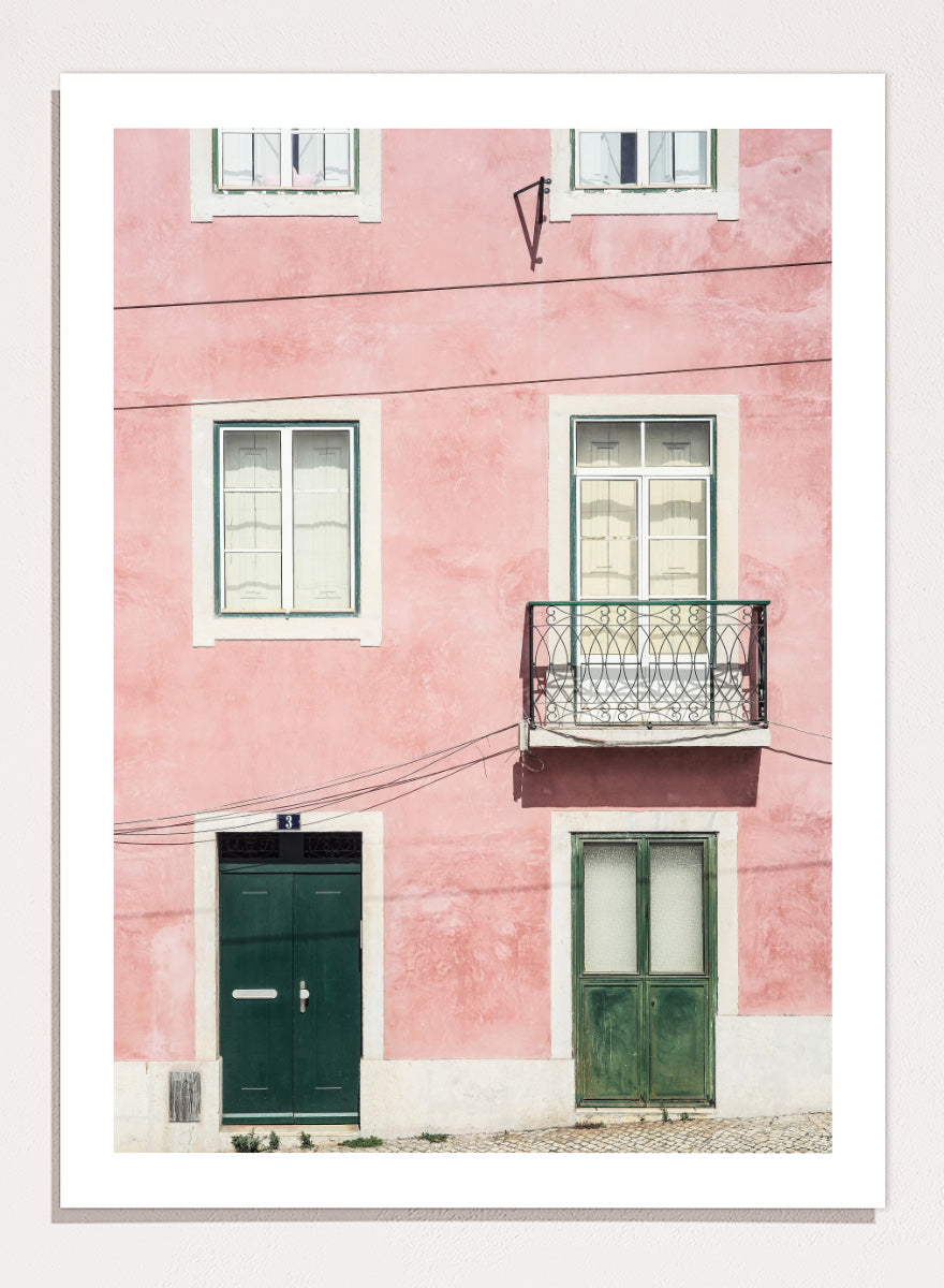 A framed art print featuring a photograph of a pink building facade with green doors, white-framed windows, and a small black metal balcony. Pink Façade Harmony by Shot by Clint architectural photography print featuring a pink exterior, green doors, white windows, and an iron balcony.