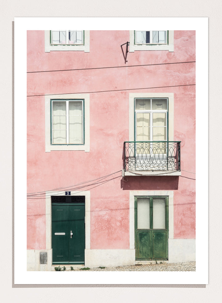 A framed art print featuring a photograph of a pink building facade with green doors, white-framed windows, and a small black metal balcony. Pink Façade Harmony by Shot by Clint architectural photography print featuring a pink exterior, green doors, white windows, and an iron balcony.