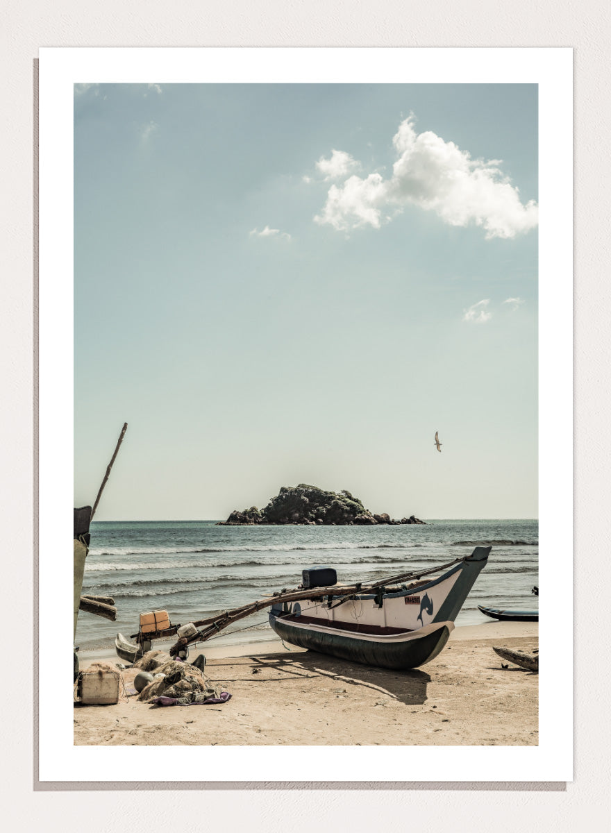 A framed photographic print of a coastal beach scene with a small boat on the sandy shore, the sea and distant rocky island, and a pale blue sky with clouds. Seaside Serenity by Shot by Clint coastal photography print featuring a fishing boat, sandy shore, ocean waves, distant island, and weathered seaside details.