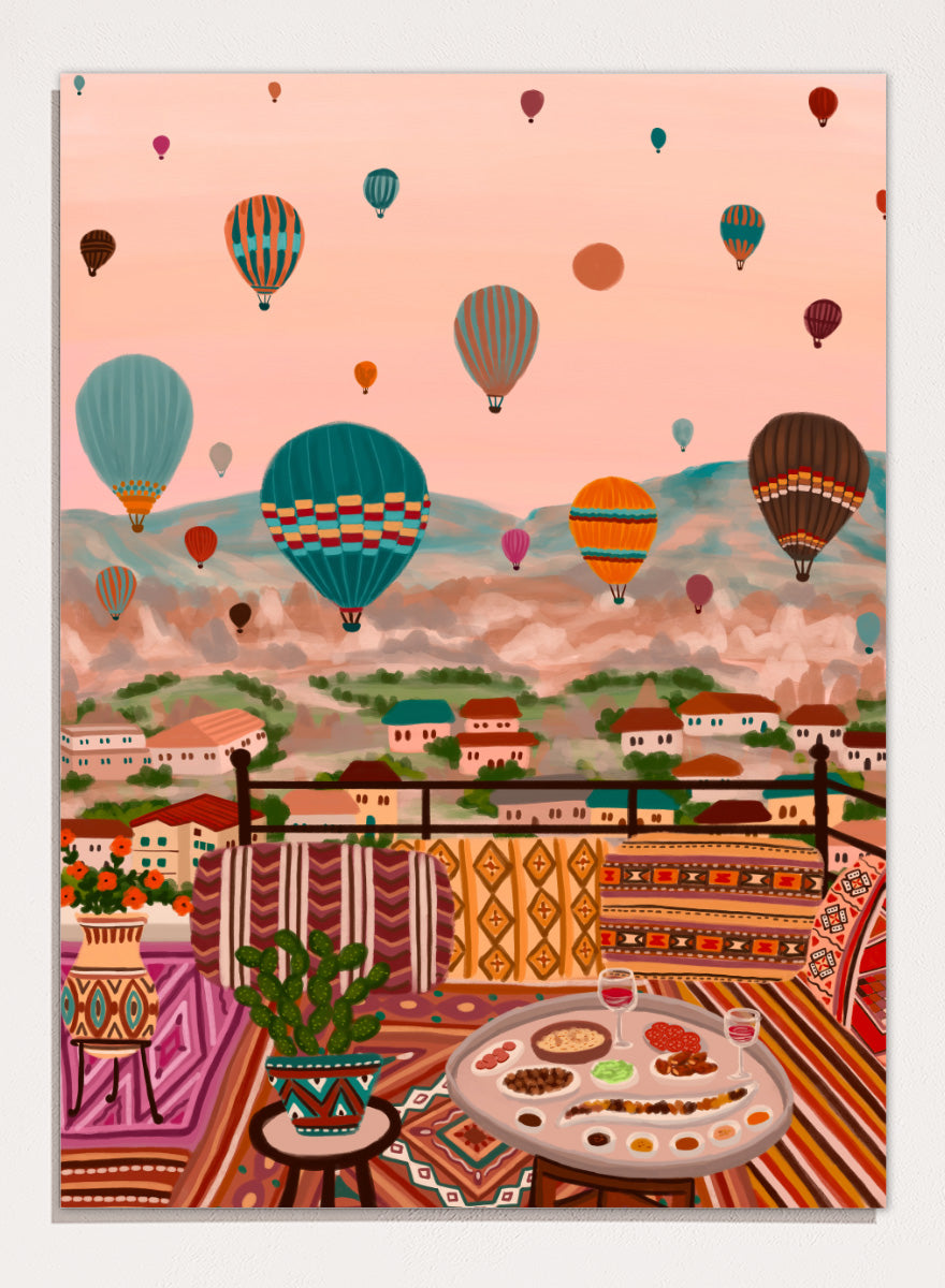 A colorful art print depicting a scenic view of a hillside town with mountains in the background, filled with hot air balloons. In the foreground, a patterned outdoor seating area features a table with food and a potted plant. Shop Balloons Over Rooftop Dinner by Nidhi Kachhadiya, a colorful travel wall art print featuring hot air balloons, rooftop dining, patterned textiles, and teal and coral tones.