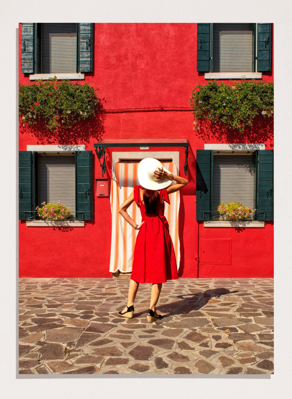 Woman in a red dress and white hat standing in front of a red wall with green shutters. Red Art Print by Anette Ohlendorf featuring a woman in a red dress standing against a red building. Officially licensed travel photography with strong color and symmetry.