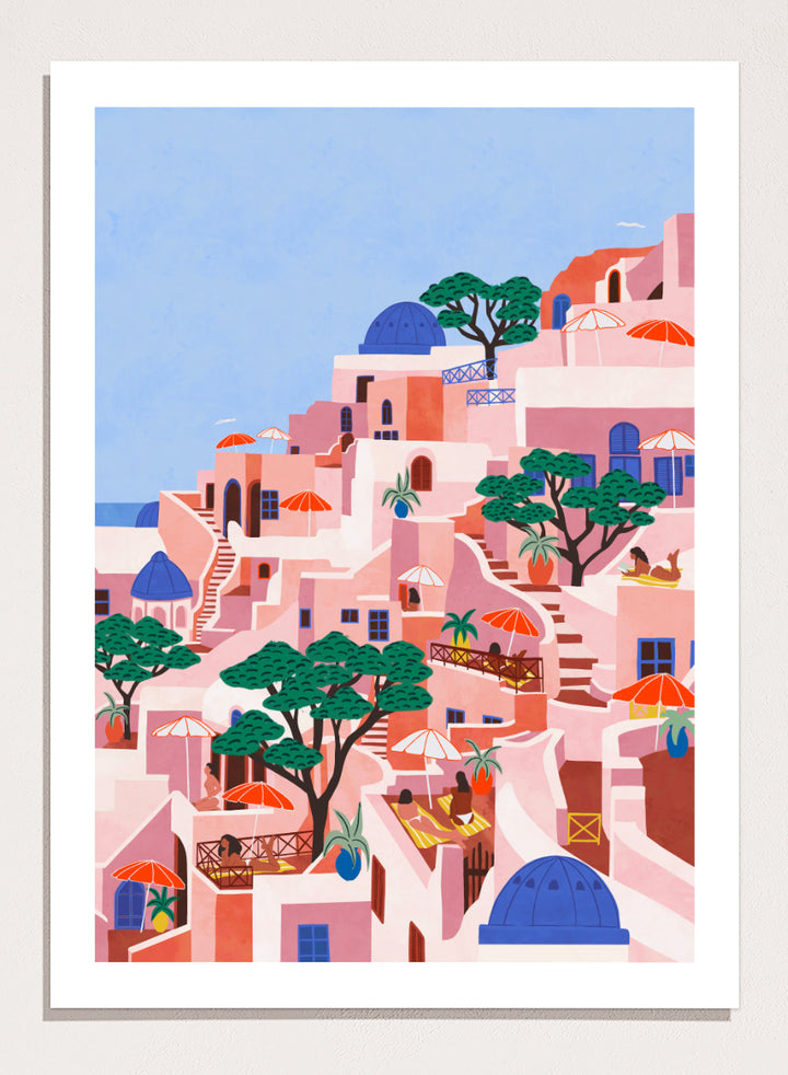 A stylized art print depicting a hillside Mediterranean village with pink and peach buildings, blue domes, green trees, and a light blue sky. Santorini Summer Dream by Ceyda Alasar art print featuring blue domes, terraced Mediterranean architecture, orange umbrellas, and sunny Greek island charm.