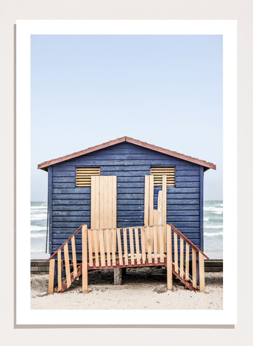 A framed photographic print of a blue wooden beach lifeguard hut on a sandy shore with the ocean and pale sky in the background. Beachside Retreat by Shot by Clint features a weathered blue beach hut and soft ocean backdrop in a calm coastal photography print.