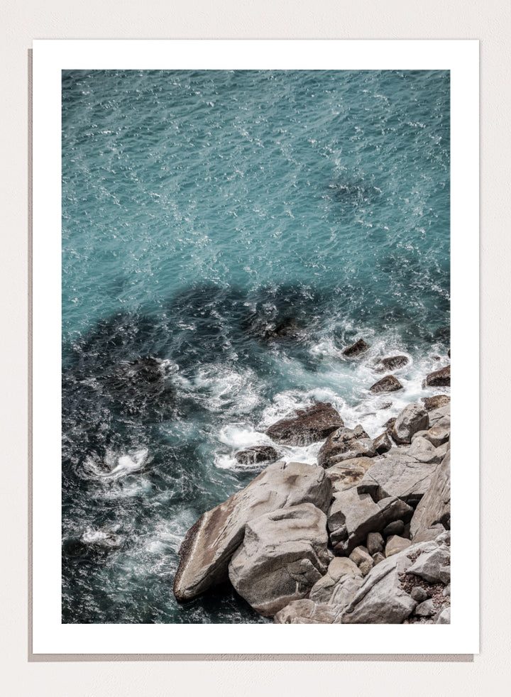 A framed photographic art print depicting a top-down view of turquoise ocean water washing over dark rocks and a rocky shoreline. Ocean’s Edge Symphony by Shot by Clint aerial ocean photography print featuring turquoise water, white surf, coastal texture, and soft seafoam tones.