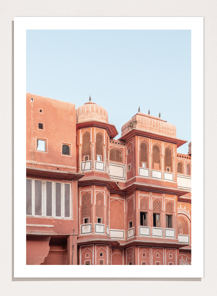 A framed art print featuring a photograph of the iconic pink sandstone Rajasthani palace, Udaipur, with white architectural details under a clear light blue sky. Terracotta Splendor by Shot by Clint architectural photography print featuring ornate palace windows, domed balconies, carved details, and warm coral tones.