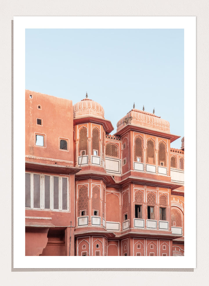 A framed art print featuring a photograph of the iconic pink sandstone Rajasthani palace, Udaipur, with white architectural details under a clear light blue sky. Terracotta Splendor by Shot by Clint architectural photography print featuring ornate palace windows, domed balconies, carved details, and warm coral tones.