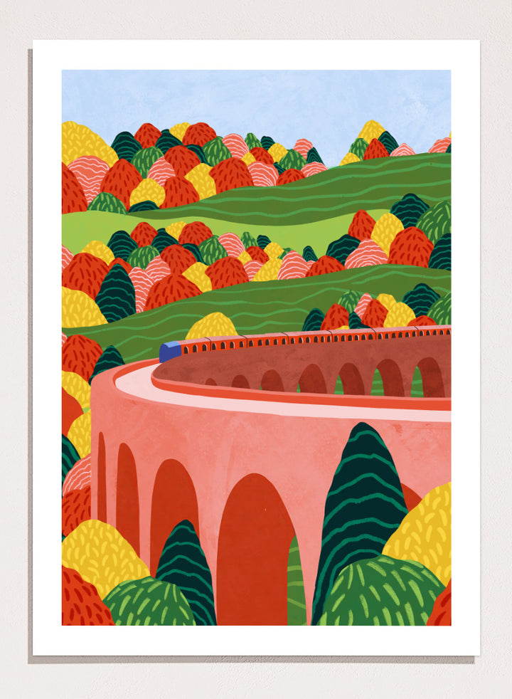 A framed art print featuring a stylized landscape illustration with rolling green hills, colorful patterned trees, and a pink arch bridge under a light blue sky. Winding Autumn Viaduct by Ceyda Alasar travel art print featuring a colorful train, arched bridge, rolling hills, and bold autumn foliage.
