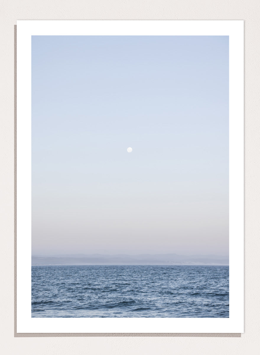 A minimalist framed art print depicting a calm blue ocean under a pale sky with a small visible moon. Moonlit Horizon by Shot by Clint coastal photography print featuring a pale moon, distant shoreline, open blue water, and soft lavender evening tones.