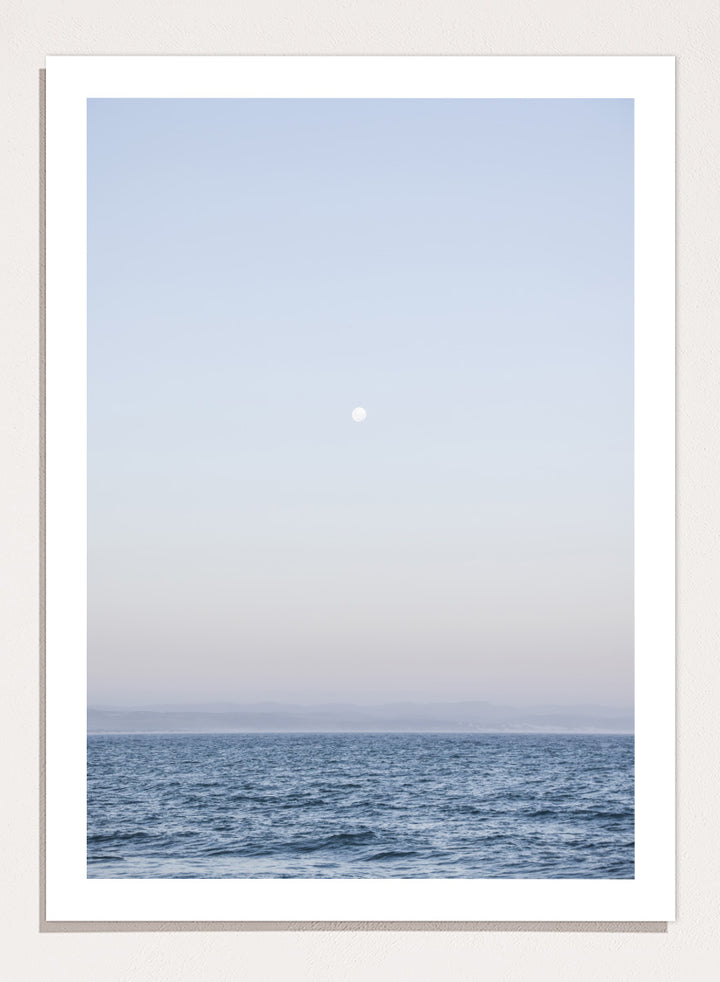 A minimalist framed art print depicting a calm blue ocean under a pale sky with a small visible moon. Moonlit Horizon by Shot by Clint coastal photography print featuring a pale moon, distant shoreline, open blue water, and soft lavender evening tones.