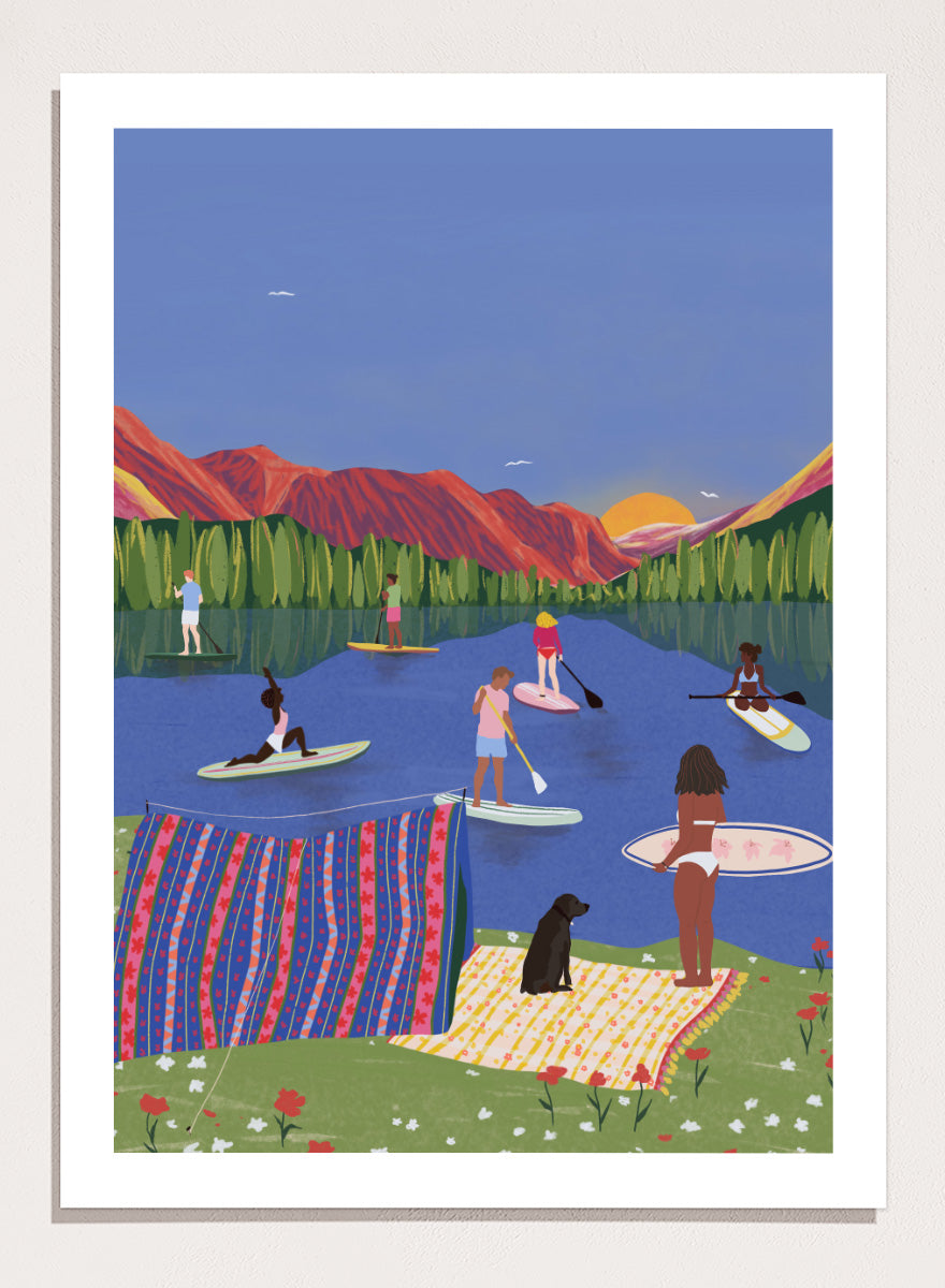 A framed art print featuring a colorful, stylized illustration of people paddleboarding and relaxing on a lake with mountains in the background. Paddleboard Lake Life by Ceyda Alasar outdoor art print featuring paddleboarders, mountain scenery, wildflowers, picnic blankets, and a lakeside dog.