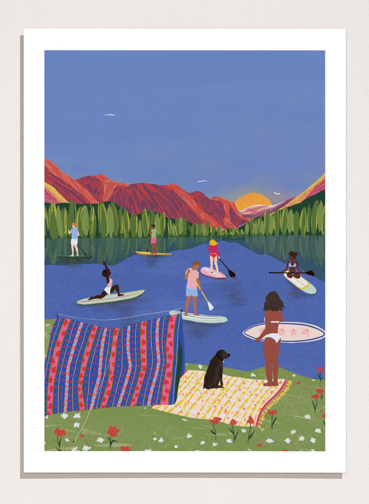 A framed art print featuring a colorful, stylized illustration of people paddleboarding and relaxing on a lake with mountains in the background. Paddleboard Lake Life by Ceyda Alasar outdoor art print featuring paddleboarders, mountain scenery, wildflowers, picnic blankets, and a lakeside dog.
