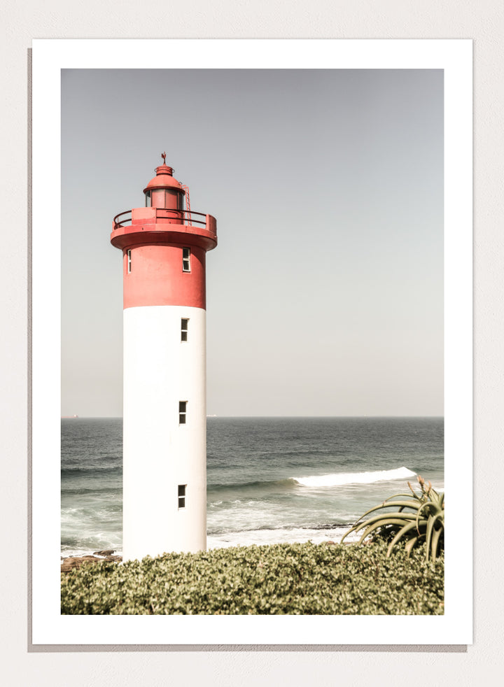 A framed photographic art print of a red and white lighthouse standing on a grassy shore with the ocean and sky in the background. Beacon by the Sea by Shot by Clint coastal photography print featuring a red-and-white lighthouse, ocean waves, pale sky, and coastal greenery.
