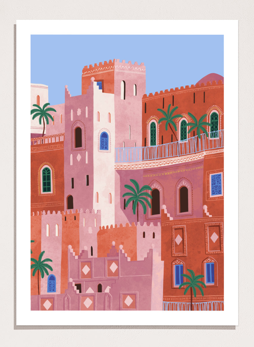 A framed art print featuring a stylized illustration of Moroccan-style buildings in shades of terracotta, pink, and cream, accented with green palm trees and blue window details, set against a light blue sky. Vibrant Moroccan City by Ceyda Alasar travel art print featuring layered terracotta architecture, arched windows, palms, and colorful Moroccan details.