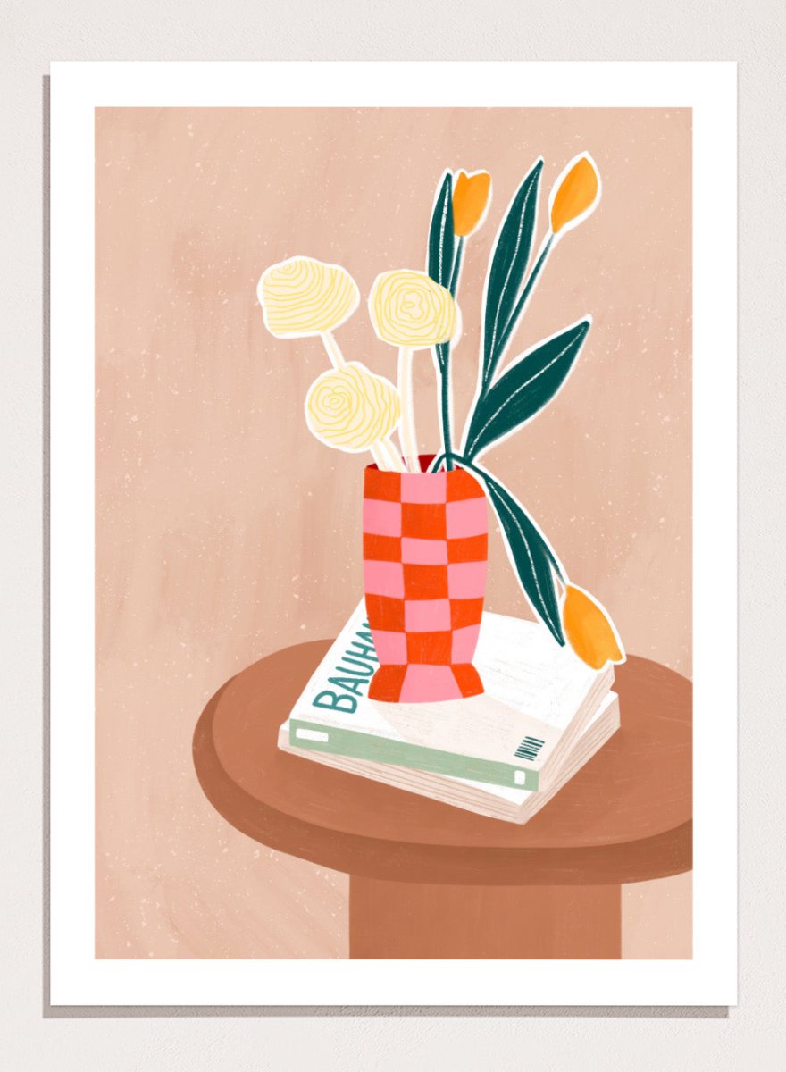 A framed minimalist art print featuring a stylized illustration of a pink and orange checkered vase filled with yellow and orange tulips, resting on a brown round table next to a white book. Colorful Tabletop Flowers by Bea Müller premium framed fine art print featuring yellow tulips and cream blooms in a red-and-pink checkered vase.