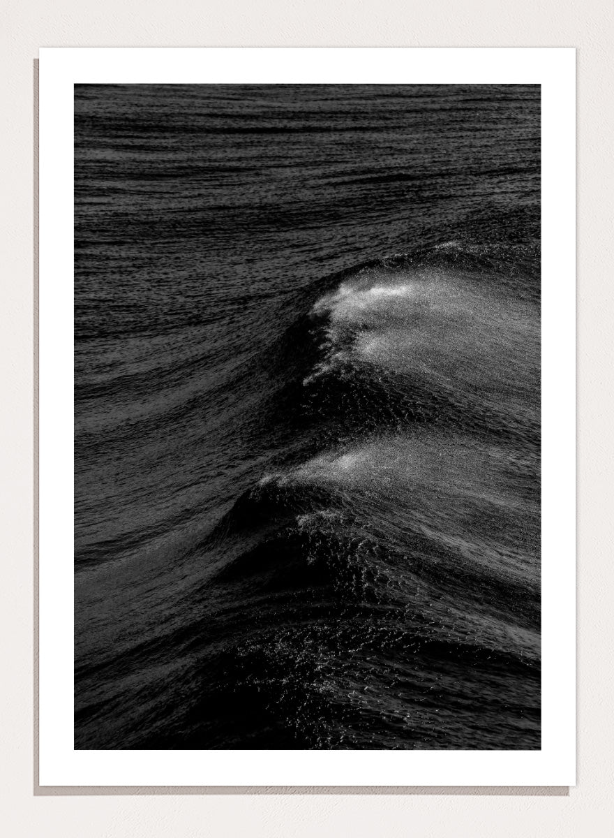 Black and white fine art photographic print of ocean waves, featuring a dramatic wave crest with white foam against dark textured water. Night Wave Cascade by Shot by Clint black-and-white ocean photography print featuring dark rolling waves, sea spray, and dramatic natural movement.