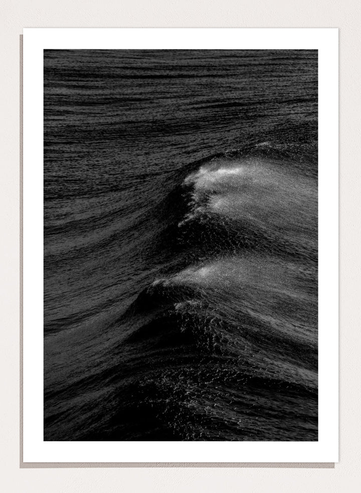 Black and white fine art photographic print of ocean waves, featuring a dramatic wave crest with white foam against dark textured water. Night Wave Cascade by Shot by Clint black-and-white ocean photography print featuring dark rolling waves, sea spray, and dramatic natural movement.