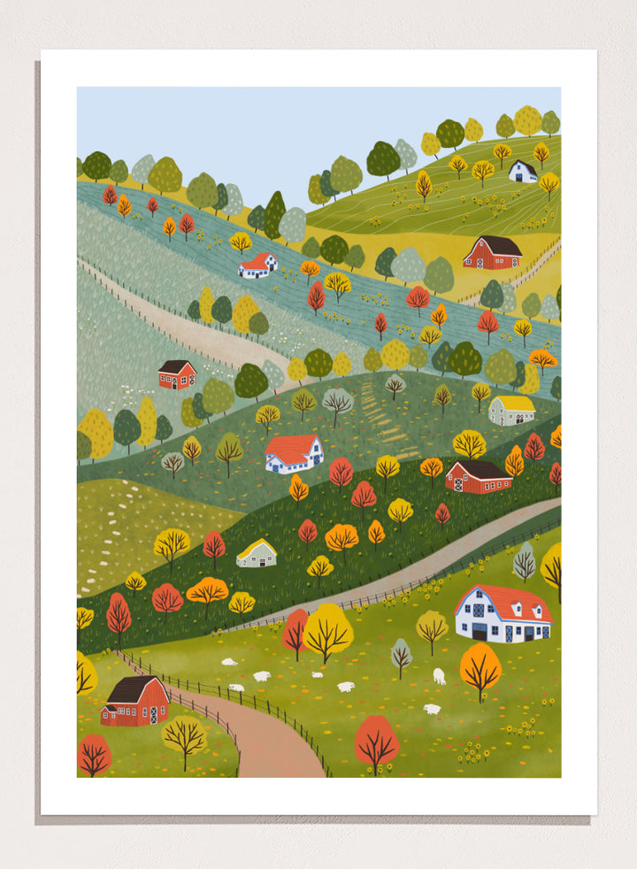 A framed art print featuring a whimsical, stylized illustration of a rolling green and yellow countryside landscape with red houses, colorful autumn trees, winding roads, and a pale blue sky. Vibrant Autumn Village Hills by Ceyda Alasar art print with rolling countryside, colorful fall trees, farmhouses, barns, sheep, and winding roads.