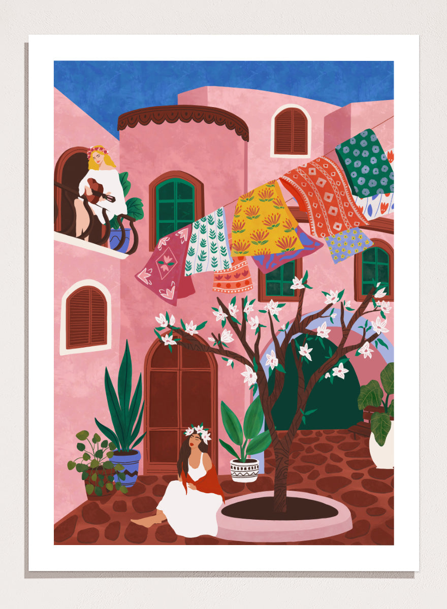 A framed art print featuring a colorful Mediterranean-style illustration of a pink courtyard with a woman sitting under a flowering tree, laundry hanging in the background, and a blue sky above. Pink Village Courtyard by Ceyda Alasar art print featuring pink architecture, patterned rugs, flowering trees, potted plants, and relaxed courtyard life.