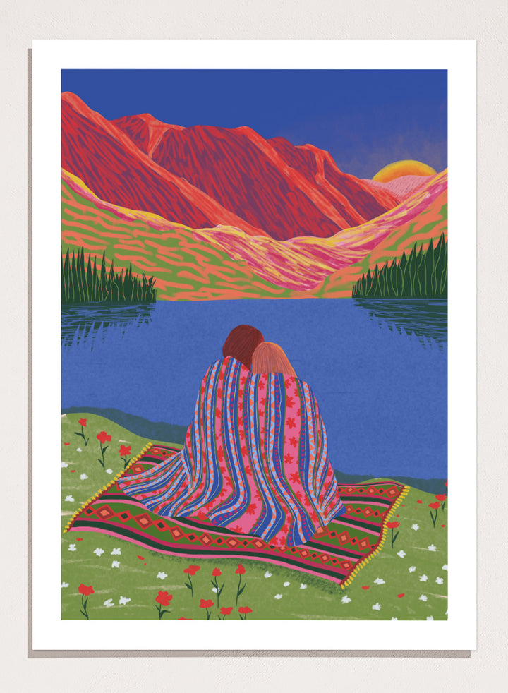 A framed art print featuring a stylized landscape illustration of two people sitting together on a patterned blanket by a blue lake, with red mountains and a yellow sun in the background. Cozy Lake Sunset art print by Ceyda Alasar features a lakeside embrace, vivid mountains, and a glowing sunset in rich, story-filled color.