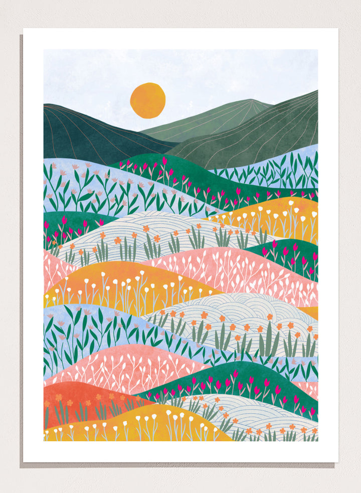 A framed art print featuring a stylized landscape illustration with rolling hills, wildflowers, mountains, and a sun in pastel and bright colors. Colorful Flower Hills by Ceyda Alasar botanical landscape art print featuring layered wildflower fields, rolling hills, mountains, and a golden sun.
