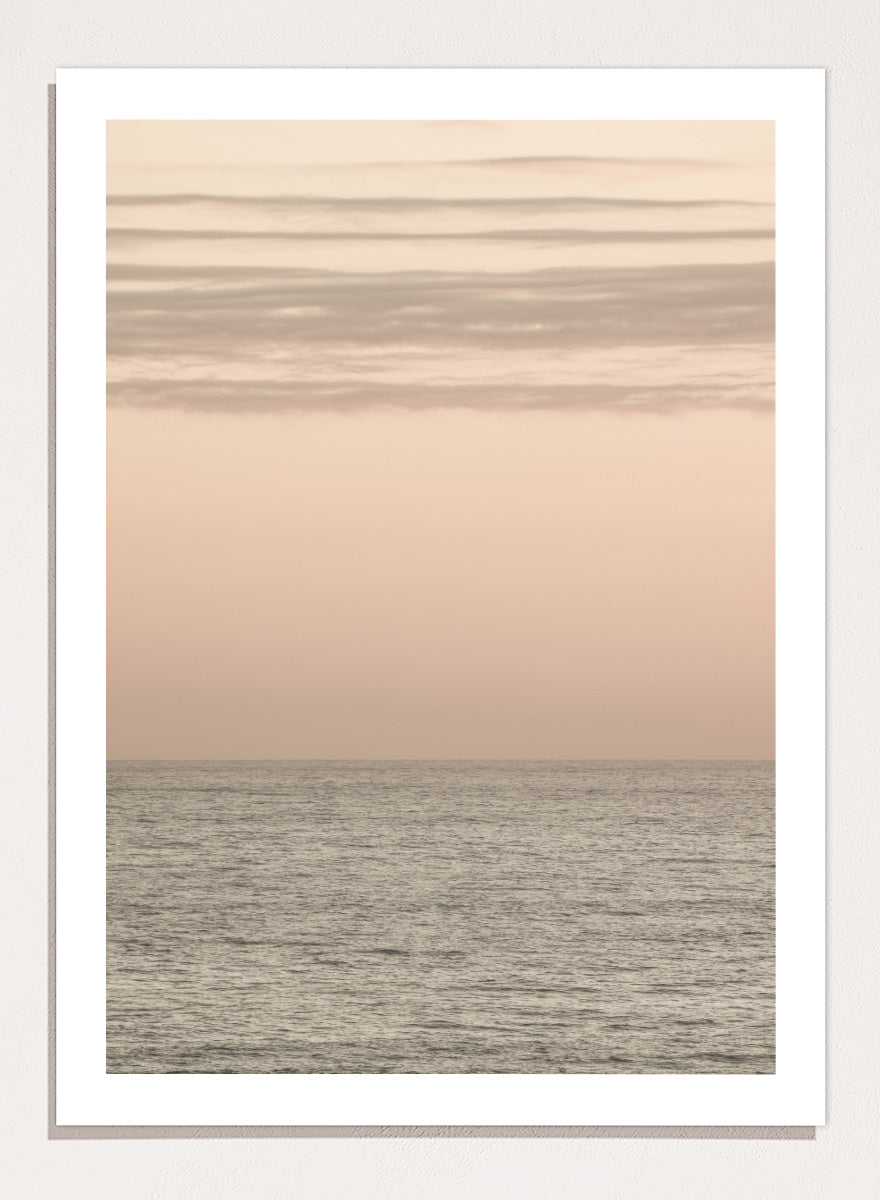 A minimalist framed art print depicting a calm seascape at dusk or dawn, with a soft peach and beige sky meeting a muted gray-blue ocean horizon. Serene Horizons by Shot by Clint features a soft peach sky and calm ocean horizon in a minimal coastal photograph with airy neutral tones.