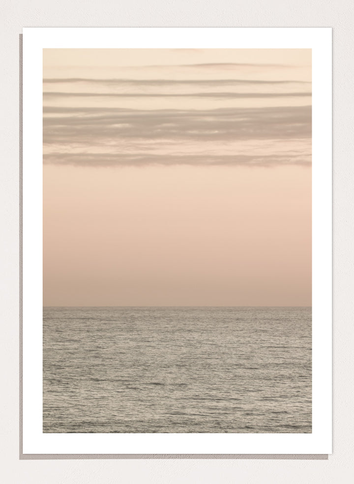 A minimalist framed art print depicting a calm seascape at dusk or dawn, with a soft peach and beige sky meeting a muted gray-blue ocean horizon. Serene Horizons by Shot by Clint features a soft peach sky and calm ocean horizon in a minimal coastal photograph with airy neutral tones.