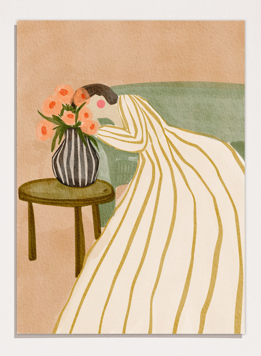 A stylized art print depicting a woman in a cream and gold striped gown leaning over a small table with a striped vase of pink flowers, set against a muted green sofa and beige background. Shop Woman Admiring Flowers by Nikita Jariwala, a modern floral wall art print featuring a woman, peach blooms, striped dress, and warm earthy tones.