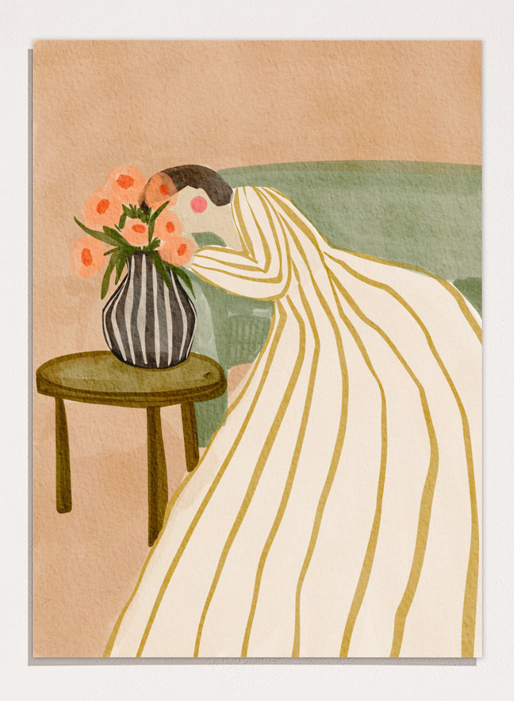 A stylized art print depicting a woman in a cream and gold striped gown leaning over a small table with a striped vase of pink flowers, set against a muted green sofa and beige background. Shop Woman Admiring Flowers by Nikita Jariwala, a modern floral wall art print featuring a woman, peach blooms, striped dress, and warm earthy tones.