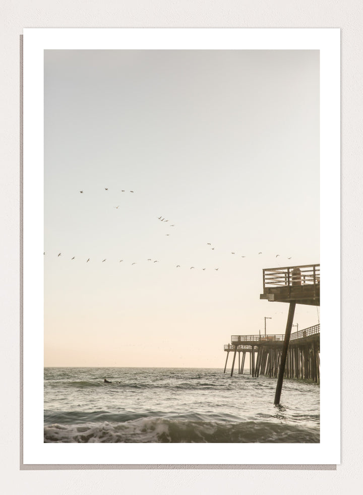 A framed photographic art print depicting a pier extending over a calm ocean under a pale sky, with birds flying across the scene. Seaside Serenity by Shot by Clint coastal photography print featuring ocean waves, a weathered pier, flying birds, and soft sunlit tones.
