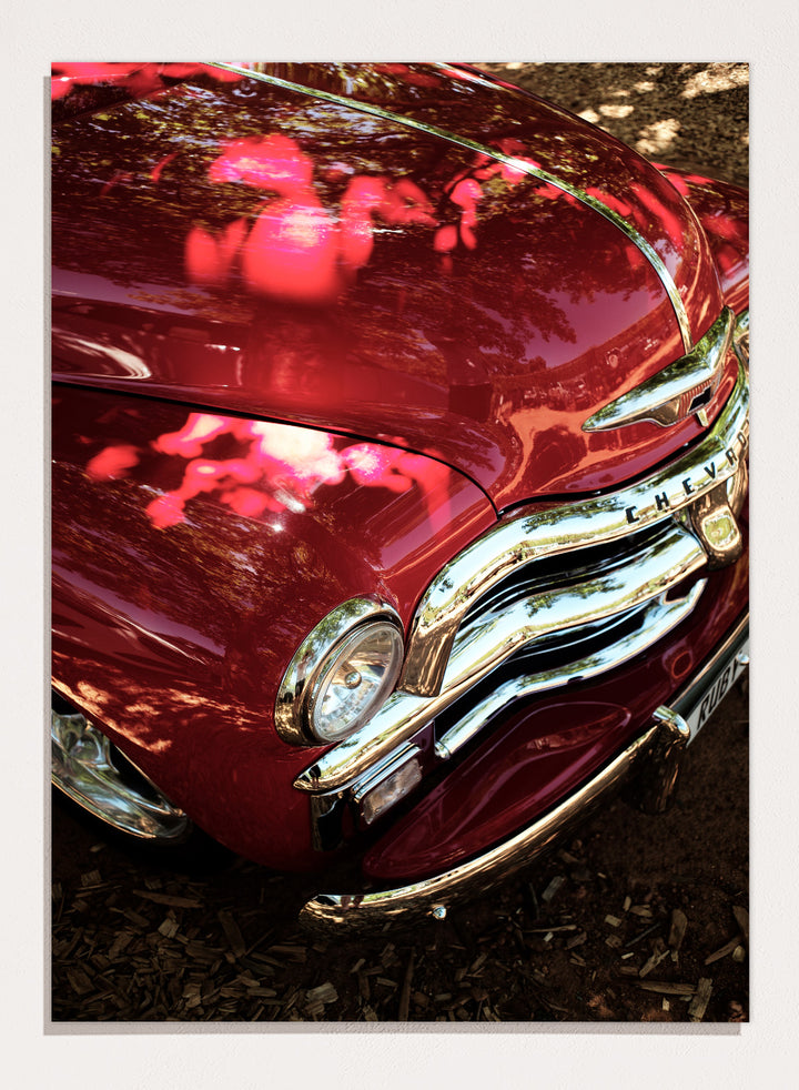 Close-up of a vintage red Chevrolet car body with chrome grille and reflections of trees on the glossy paint, photographed outdoors on mulch. Vintage Red Car by Shot by Clint automotive photography print featuring a classic Chevrolet, deep red paint, polished chrome, and sculptural vintage details.