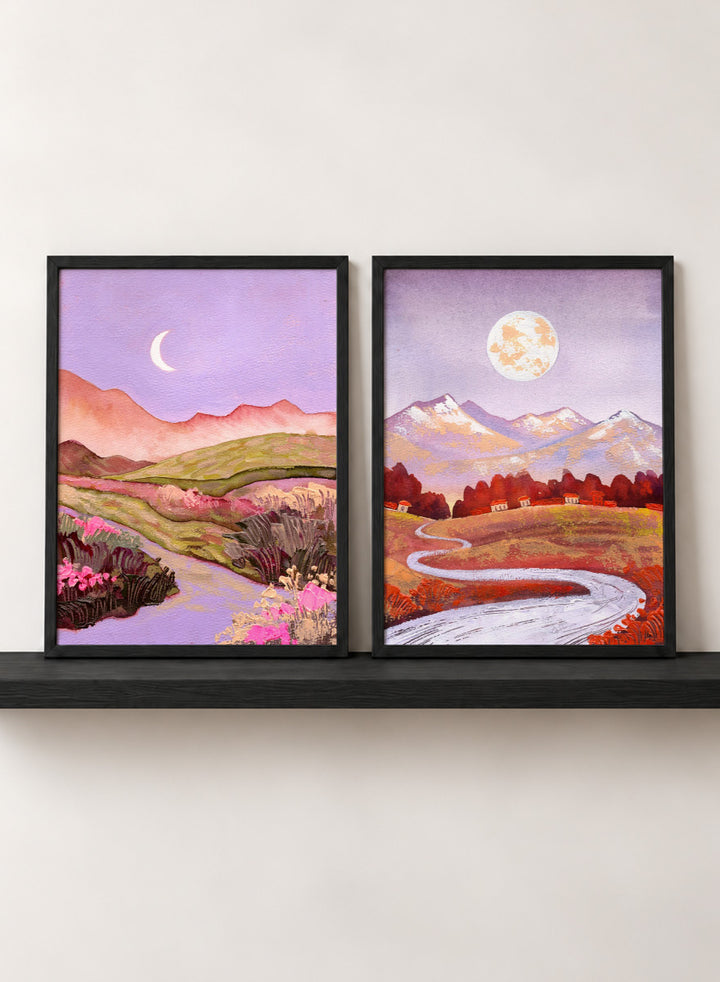 Two framed landscape art prints displayed side by side on a black shelf against a light beige wall. The left print shows a purple-toned valley with a crescent moon, pink flowers, and distant hills. Shop Moonlit Landscapes by Ekaterina Prisich, a set of 2 landscape wall art prints posters featuring moonlit mountains, winding paths, and purple tones.