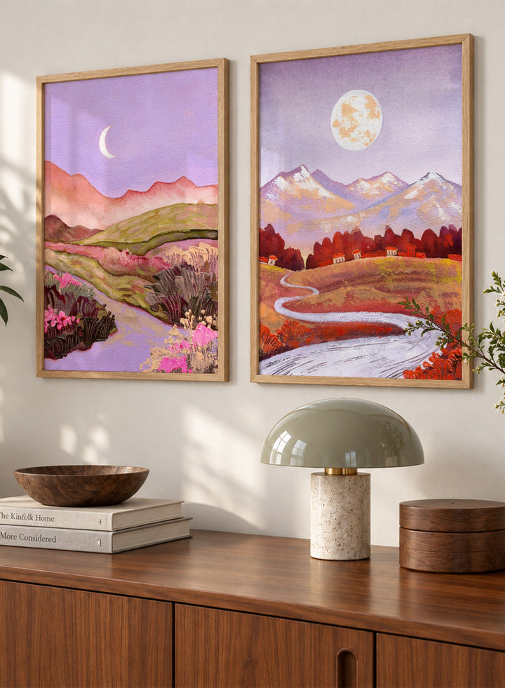 Two framed landscape art prints displayed side by side on a black shelf against a light beige wall. The left print shows a purple-toned valley with a crescent moon, pink flowers, and distant hills. Shop Moonlit Landscapes by Ekaterina Prisich, a set of 2 landscape wall art prints posters featuring moonlit mountains, winding paths, and purple tones.