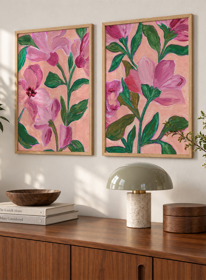 Two framed floral art prints with pink flowers and green leaves on a peach background. Shop Vibrant Pink Botanicals by Nikita Jariwala, a set of 2 floral wall art prints posters featuring oversized pink flowers and lush botanical artwork.