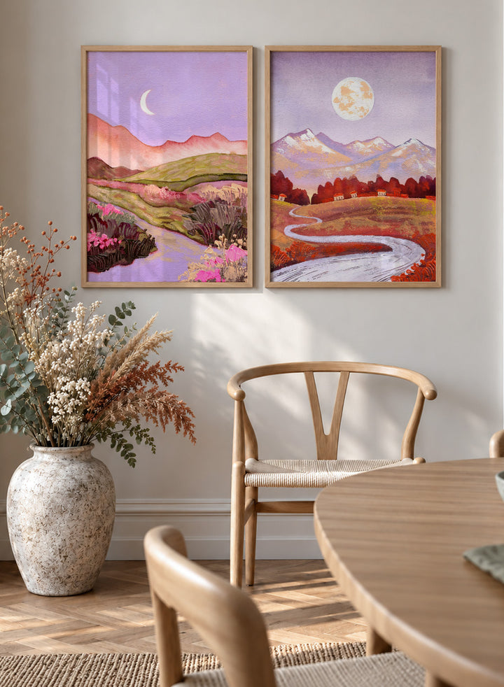 Two framed landscape art prints displayed side by side on a black shelf against a light beige wall. The left print shows a purple-toned valley with a crescent moon, pink flowers, and distant hills. Shop Moonlit Landscapes by Ekaterina Prisich, a set of 2 landscape wall art prints posters featuring moonlit mountains, winding paths, and purple tones.