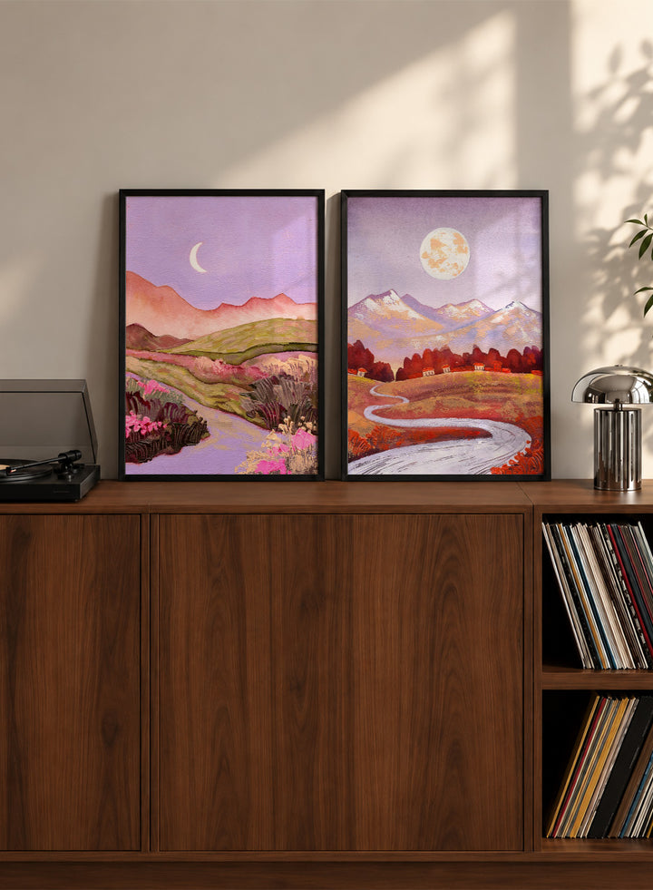 Two framed landscape art prints displayed side by side on a black shelf against a light beige wall. The left print shows a purple-toned valley with a crescent moon, pink flowers, and distant hills. Shop Moonlit Landscapes by Ekaterina Prisich, a set of 2 landscape wall art prints posters featuring moonlit mountains, winding paths, and purple tones.