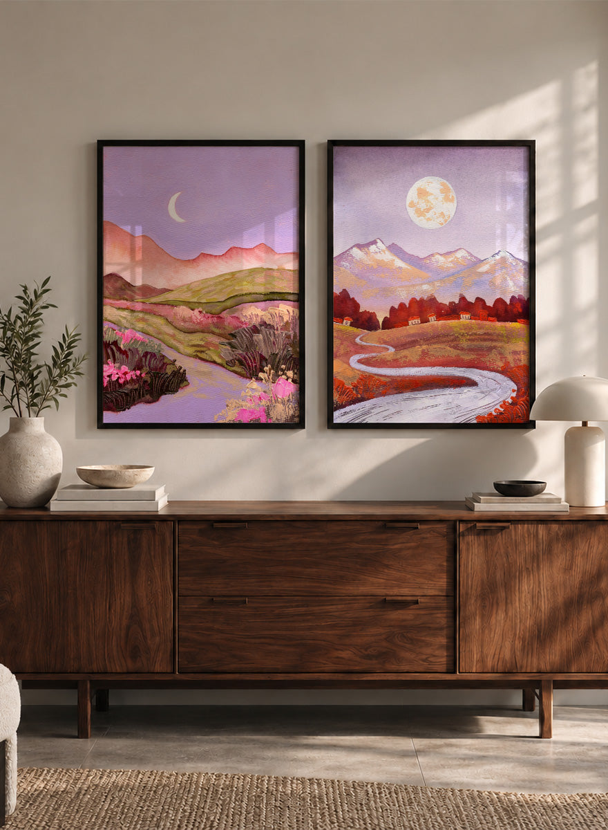 Two framed landscape art prints displayed side by side on a black shelf against a light beige wall. The left print shows a purple-toned valley with a crescent moon, pink flowers, and distant hills. Shop Moonlit Landscapes by Ekaterina Prisich, a set of 2 landscape wall art prints posters featuring moonlit mountains, winding paths, and purple tones.