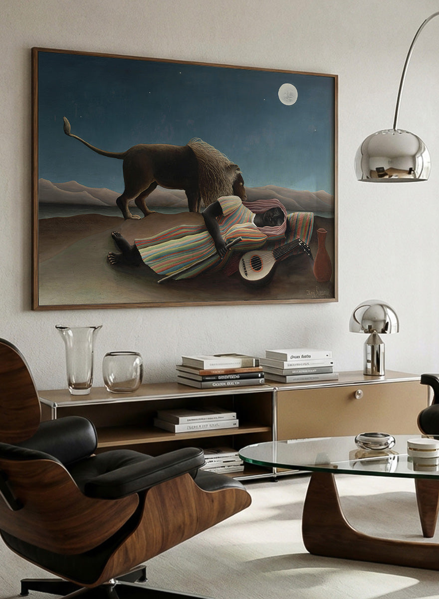 A framed surreal art print depicting a lion standing over a sleeping person wrapped in a striped blanket, with a guitar, vase, and full moon in a night sky. The Sleeping Gypsy by Henri Rousseau is a vintage art print featuring a sleeping traveler, lion, moonlit desert, striped robe, mandolin, and deep blue night sky.