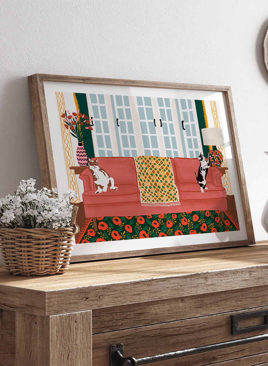 A framed art print depicting a cozy living room scene with a coral-pink sofa, two cats, a patterned throw blanket, and a floral rug. Playful Cats Living Room by Ceyda Alasar cat art print featuring two mischievous cats, a coral sofa, floral rug, patterned throw, and colorful decor.