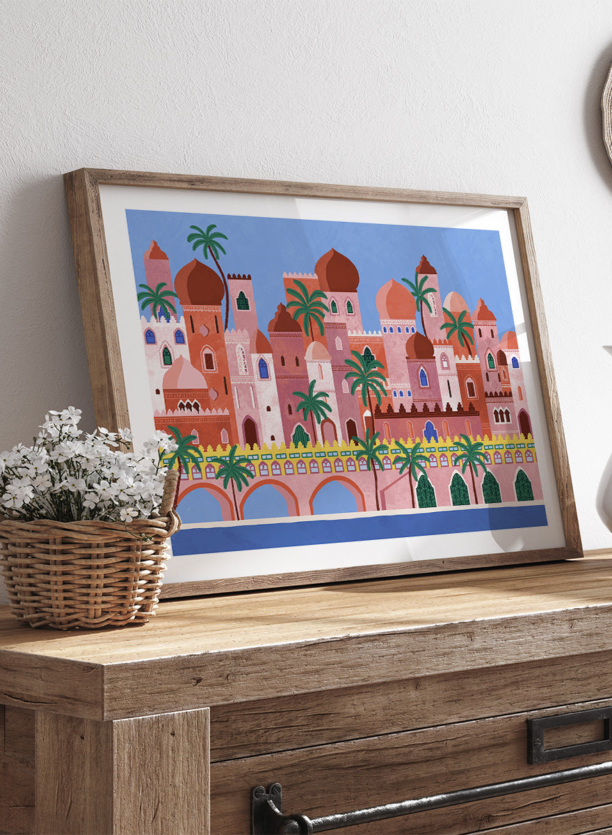 A framed art print featuring a stylized illustration of a coastal city with pink and peach buildings, green palm trees, and a blue sky. Arabian Fantasy City by Ceyda Alasar travel art print featuring ornate domes, arches, palm trees, patterned facades, and colorful desert architecture.