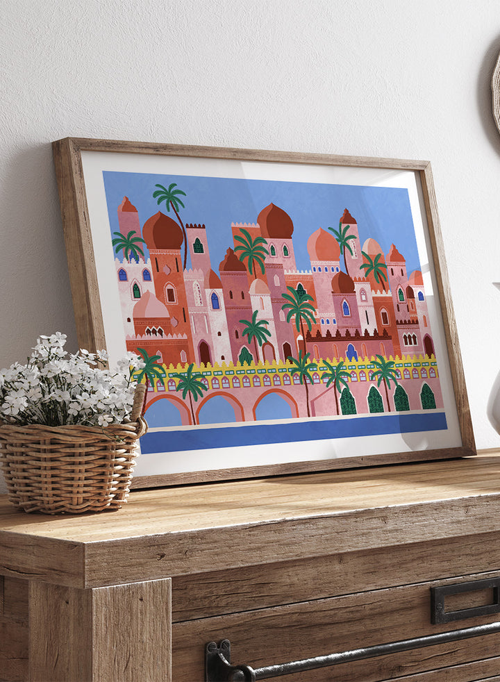A framed art print featuring a stylized illustration of a coastal city with pink and peach buildings, green palm trees, and a blue sky. Arabian Fantasy City by Ceyda Alasar travel art print featuring ornate domes, arches, palm trees, patterned facades, and colorful desert architecture.