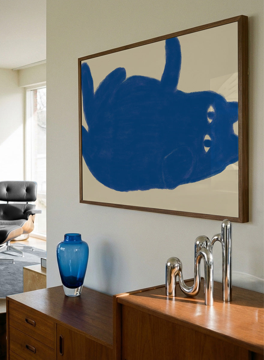 A framed art print featuring a bold blue abstract cat illustration on a warm beige background. Whimsical Blue Cat by The Art Concept is a playful cat art print featuring a lounging blue cat on a warm cream background in a bold minimalist style.