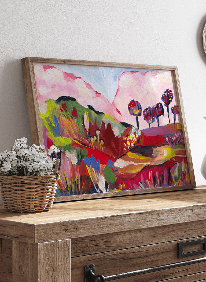 A framed abstract landscape painting featuring pink clouds, green hills, and colorful stylized trees. Shop Vibrant Fantasy Landscape by Dirkie Nell, a colorful landscape art print featuring pink clouds, rolling hills, wildflowers, and bold abstract color.