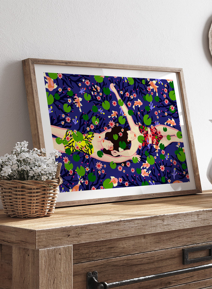A framed art print featuring a stylized illustration of a woman lying on a dark blue background covered with green lily pads and pink flowers. Serene Night Float by Ceyda Alasar friendship art print featuring three women floating among lily pads, flowers, and fish in a deep blue pond.