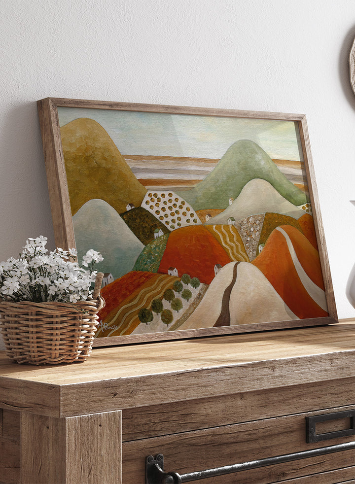 A framed landscape painting featuring stylized rolling hills and fields in earthy tones of orange, green, beige, and brown. Shop Colorful Hillside Village by Angeles M. Pomata, an autumn landscape art print featuring rolling hills, tiny homes, winding roads, and warm earthy tones.