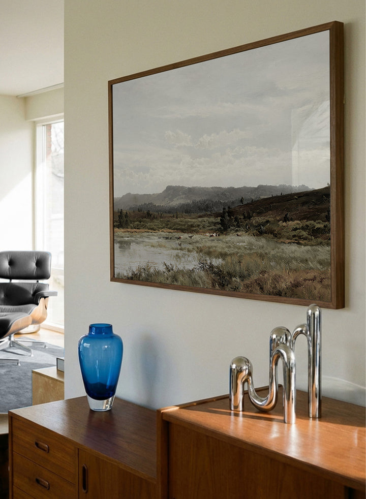 A framed landscape art print depicting a muted, overcast valley scene with a small body of water, grassy foreground, distant hills, and a cloudy sky, shown against a light neutral background. A View of Fleet Moor, Surrey by Alfred de Bréanski is a vintage English landscape print featuring cattle, water, rolling hills, open countryside, and a cloudy sky.