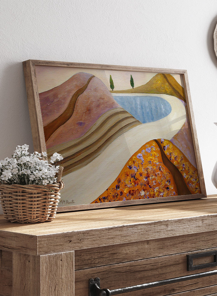 A framed landscape painting featuring stylized hills, a winding cream path, a blue pool, and two small green trees. Shop Peaceful Hills and Lake by Angeles M. Pomata, a serene landscape art print featuring a blue lake, rounded hills, winding paths, and soft earthy tones.