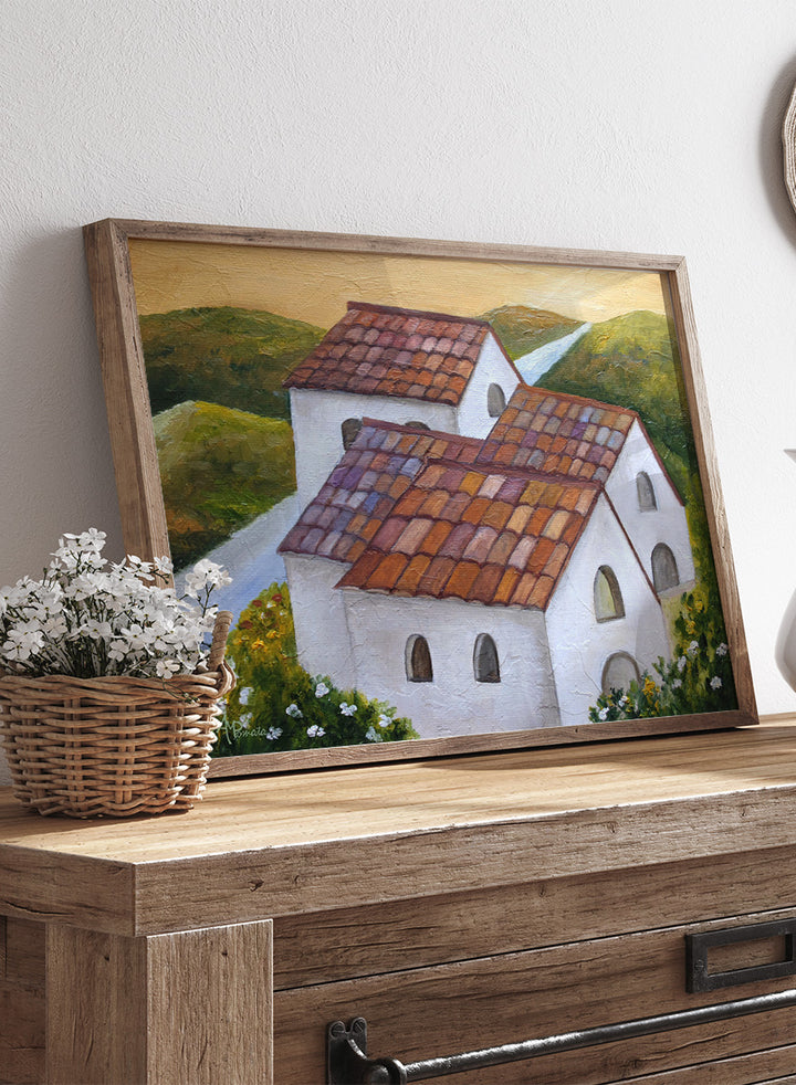 A framed landscape painting depicting two white buildings with terracotta tiled roofs nestled in green hills, with a winding light blue road and a warm yellow sky. Shop Tuscan Hillside Village by Angeles M. Pomata, a warm countryside landscape art print featuring white houses, terracotta roofs, green hills, and wildflowers.