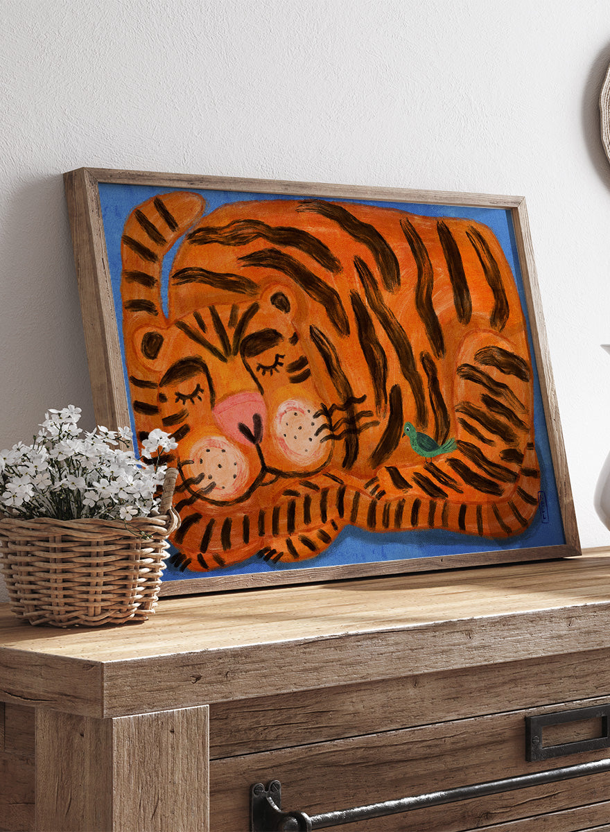 A framed painting of a sleeping tiger on a blue background. The tiger is orange with bold black stripes, has a pink nose and cheeks, and a small green bird near its side. A framed painting of a sleeping tiger on a blue background. The tiger is orange with bold black stripes, has a pink nose and cheeks, and a small green bird near its side. 